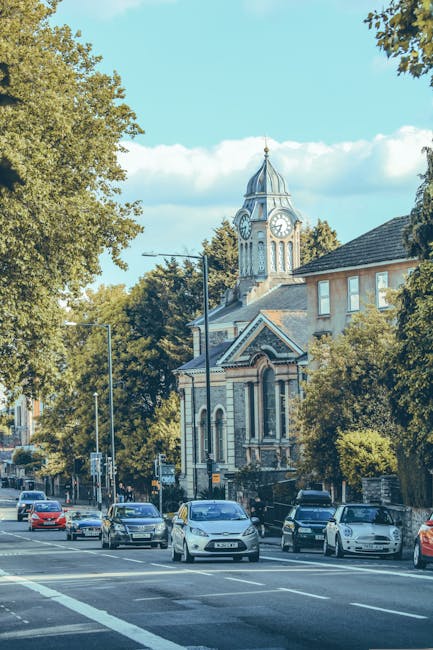 A view of a street in Kentish Town during daytime with several parked cars along the pavement and moving vehicles on the road. In the background, there is a historic clock tower with a spire, surrounded by trees and residential buildings. The scene features clear blue skies and sunlight filtering through the leaves, indicating good weather. This setting reflects a typical environment where home relocation or furniture transport may take place, with visible urban infrastructure that accommodates loading and unloading activities often involved in professional removals by Kentish Town Removals. Although no furniture or moving actions are visible, the image captures the context of a residential neighbourhood suitable for delivery, packing, and transportation of household items in connection with removals services.