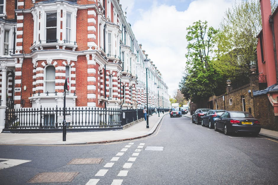 A street view in a residential area featuring a row of Victorian-style terraced houses with ornate white and red brick facades, large bay windows, and decorative trim. The street is lined with parked cars on both sides, including a black vehicle close to the foreground and several others further down the road. A few pedestrians are visible along the sidewalk, which is separated from the road by a black metal fence. The bright daylight highlights the greenery of a large tree with fresh leaves on the right side of the image, situated behind the brick wall of one property. In the background, the sky is partly cloudy with patches of blue. This scene depicts a typical urban setting suitable for home relocation and furniture transport, with open roadway and accessible parking for moving vehicles. The image aligns with house removals and packing services, such as those offered by Kentish Town Removals, and captures the environment for a professional moving process.