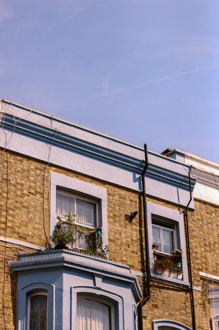 A view of a street in Kentish Town during daytime with several parked cars along the pavement and moving vehicles on the road. In the background, there is a historic clock tower with a spire, surrounded by trees and residential buildings. The scene features clear blue skies and sunlight filtering through the leaves, indicating good weather. This setting reflects a typical environment where home relocation or furniture transport may take place, with visible urban infrastructure that accommodates loading and unloading activities often involved in professional removals by Kentish Town Removals. Although no furniture or moving actions are visible, the image captures the context of a residential neighbourhood suitable for delivery, packing, and transportation of household items in connection with removals services.