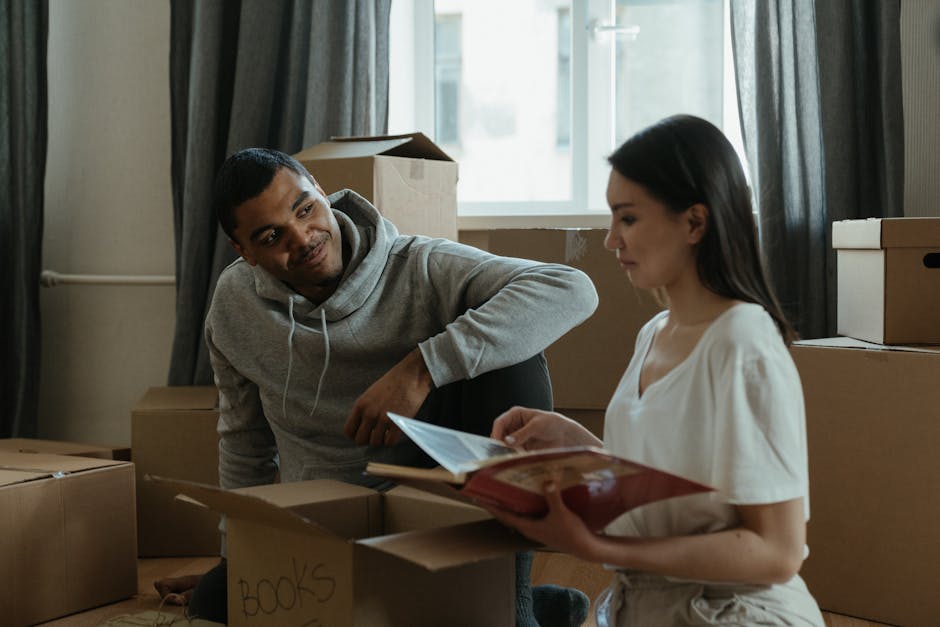 A man and a woman are inside a room filled with packed cardboard moving boxes, some labeled with handwritten tags such as 'BOOKS'. The man is kneeling on the floor, wearing a grey hoodie, and is smiling while looking at the woman. The woman, dressed in a white blouse, is standing and holding an open folder or document in her hands, reading or examining it. Behind them, a window allows natural light into the space, and dark curtains frame the window. Several large cardboard boxes are stacked around them, some open or closed, indicating a home relocation process. The scene depicts a casual moment during packing or unpacking, with visible packing materials like tape and labels, reflecting a typical house moving or removals activity, in a setting that could be part of a professional service like Kentish Town Removals.