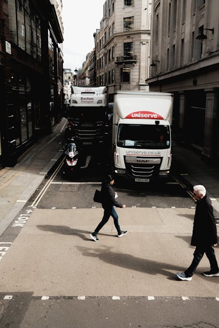 A city street intersection during daytime showing a white moving van with the logo 'uniserve' parked partly on the pavement and partially on the road, with another truck behind it. A black motorcycle is parked next to the vehicles. Two pedestrians, a woman and an older man, are crossing the road at a pedestrian crossing with white striped markings; the woman is in casual clothing and carrying a bag, while the man has gray hair and is wearing a dark suit. Surrounding the scene are tall, multi-storey buildings with a mix of modern and historic architectural styles, and the sky appears partly cloudy. The image illustrates urban moving logistics, with vehicles connected to house removals and furniture transport, as well as the process of packing and preparing for a home relocation, in the context of services provided by Kentish Town Removals.