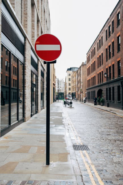 A close-up view of a red and white 'no entry' traffic sign mounted on a black pole placed on a paved sidewalk next to a narrow street in an urban area. The street features brick and stone buildings with large windows on both sides, some of which have reflective glass. In the distance, there are a few pedestrians walking along the sidewalk and bicycles parked near the curb. The scene is taken in natural daylight with an overcast sky, and the street has a slightly wet appearance, possibly from recent rain. This setting is typical of an urban environment suitable for house removals or furniture transport, with the street layout and building facades visible for context. Kentish Town Removals may operate in similar environments, particularly where narrow access is a challenge for home relocation services.