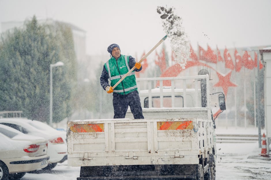 A man dressed in a green high-visibility vest, dark trousers, and a blue beanie hat is standing on the bed of a white flatbed truck during a snowstorm, while shovelling snow with a long-handled snow shovel. Snow is falling heavily around him, and he appears to be clearing the area for house relocation or moving purposes. The background includes parked cars covered with snow, a building with a colorful graffiti mural, and an overcast sky, indicating winter weather conditions. The scene captures the logistics and preparation involved in a house removal service, with snow prevention and vehicle access measures being a priority. Kentish Town Removals' trusted team is engaged in managing transport logistics in challenging weather, ensuring safe furniture transport and packing for home relocation, aligning with professional moving services at [PAGE_TITLE] on [PAGE_URL].