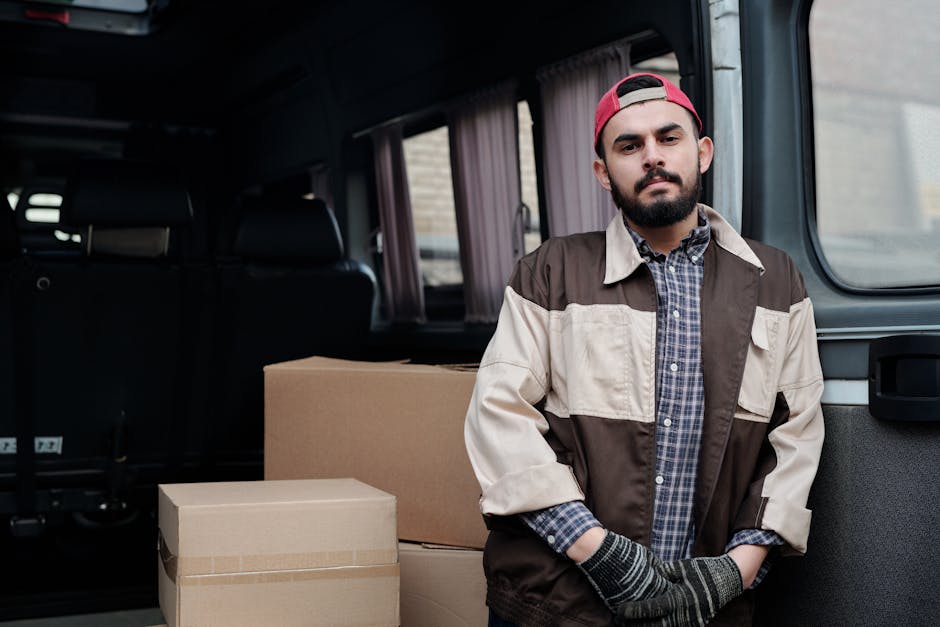 A man with a beard, wearing a red cap backwards, a plaid shirt, and a beige and brown jacket, stands beside the open rear door of a grey van during a house relocation. Inside the van, there are several stacked cardboard boxes of various sizes, some wrapped in protective plastic or packing tape, arranged neatly for transport. The vehicle is parked outdoors on a paved area, with a brick building visible through the van's windows. The scene depicts a loading process typical of furniture transport and packing and moving services, with the man likely involved in overseeing or assisting with the loading of household items. The lighting is natural, and the focus is on the man and the boxes, highlighting the logistics involved in small flat removals as offered by Kentish Town Removals, which specialises in home relocation services.