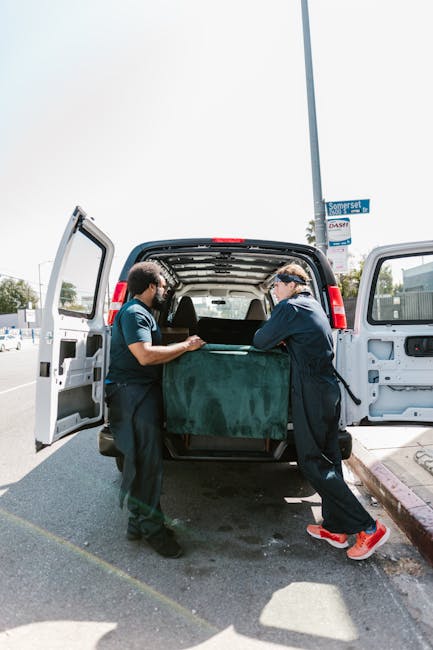 Two movers from Kentish Town Removals are seen loading a large, dark green upholstered piece of furniture into the back of an open white van on a street. The furniture is partially inside the vehicle, with one mover holding and guiding it, while the other assists from the side. The van is parked on a paved road, adjacent to a sidewalk with a lamppost marked 'Somerset,' and other vehicles and buildings are visible in the background. The interior of the van reveals a spacious cargo area prepared for home relocation or furniture transport, with the open rear doors allowing easy access for loading. The scene captures the process of packing and moving household items, typical of small flat removals, with the movers wearing casual work attire, including dark Protective clothing and gloves to facilitate safe handling during the loading process. This activity reflects professional moving services provided by Kentish Town Removals, ensuring efficient packing and transportation within the context of house removals.