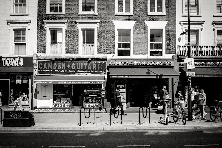 A black-and-white image depicts a city street scene outside a building with multiple retail storefronts, including a music shop and a bookstore, with signage indicating the businesses' names. The building has three floors, with large windows on each level. The ground floor storefronts are accessed via pavement, where a group of people is seen walking along the sidewalk, some engaged in conversation or carrying belongings. Several bicycles are parked along the curb, secured to metal bike racks, and there is a streetlamp near the sidewalk’s edge. The scene includes a vehicle parked close to the shops, and a few pedestrians are positioned near the storefronts, possibly observing or waiting. This setting exemplifies urban activity typical of a busy local shopping area, suitable for a commercial or residential move, reflecting moving services that involve furniture transport, packing, and loading processes associated with house removals in Camden. Kentish Town Removals often coordinate such tasks in similar environments to facilitate home relocation or furniture transport within dense urban areas.