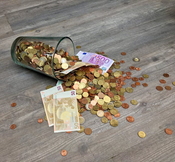 A clear glass jar tipped over on a wooden floor with its contents of euro coins and banknotes spilled out. The coins include various denominations such as 1, 2, and 5 euro cents, as well as higher-value coins, scattered across the floor. Several 50 euro banknotes are laid flat on the floor among the coins, with others partially visible underneath. The scene appears to depict a large sum of money spilled, with the coins and notes spread out around the jar. This image relates to financial aspects of house removals and moving costs, as presented on Kentish Town Removals' website, which discusses saving money through various moving options.
