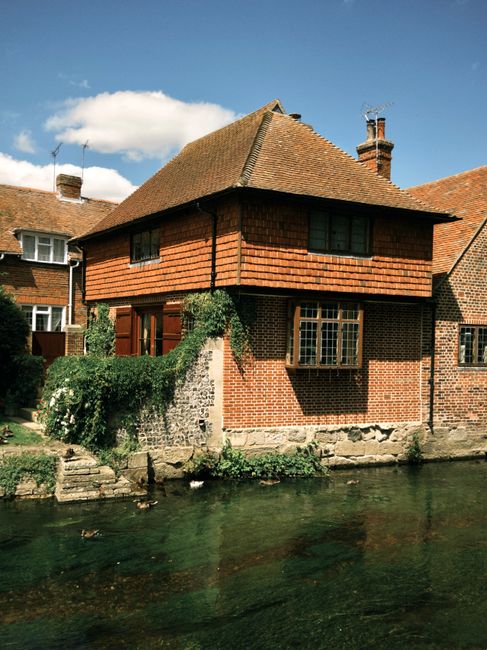 A daytime view of a two-storey brick house situated beside a narrow river or canal, with the lower part of the building resting on a stone foundation. The house features a combination of brickwork and wooden shingle cladding on the upper level. There are multiple windows, including a bay window on the ground floor and smaller windows on the upper floor. A chimney is visible on the roof, which is covered with brown tiles. Garden vegetation, including bushes and climbing plants, partially surrounds the house, some growing over the low stone wall that separates the property from the water. In the foreground, the river's clear water reflects the house and sky, with a few ripples visible on the surface. The sky above is partly cloudy with a bright blue backdrop, indicating good weather. This setting illustrates a typical residential scene that might involve home relocation or moving activities, with the house close to the water and prepared for packing and transport, as often facilitated by Kentish Town Removals.