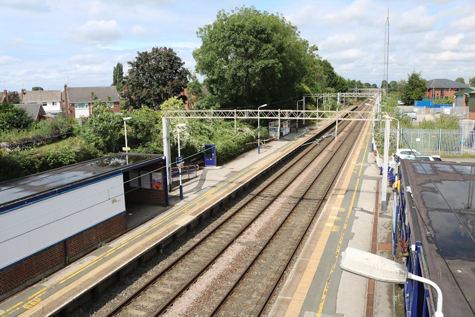A high-angle view of a suburban train station platform during daytime, with two parallel tracks running through the centre. On the left side of the image, there is a small brick building with a blue-tiled roof, covered by a black canopy, and a white sign attached to the wall. The platform features yellow tactile paving along the edge for safety, with a blue ticket machine and a small waiting area. On the right side, a black bus with reflective windows is partially visible, parked close to the platform. Overhead, a white metal gantry supports overhead electric lines running alongside the tracks, with electrical insulators and support wires. In the background, lush green trees and residential houses with sloped roofs can be seen, along with a taller communication tower just beyond the station. The scene is well-lit under a partly cloudy sky, and the environment reflects a typical urban railway station setting, appropriate for discussing home relocation transport or moving logistics as handled by Kentish Town Removals.