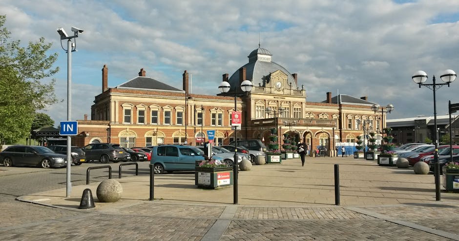 A large brick building with Victorian architectural features, including arched windows and a central clock tower, situated in a paved public square. The square has parked cars along the perimeter, decorative street lamps, and planters with flowers. A person is walking across the open space near the building entrance, which suggests activity related to home relocation or moving services. The sky is partly cloudy, with natural daylight illuminating the scene. This image captures a typical setting for house removals or furniture transport in a town centre, with the building and surrounding environment indicating proximity to a train station. Kentish Town Removals occasionally supports such moving logistics, especially in areas near busy stations like Kentish Town.