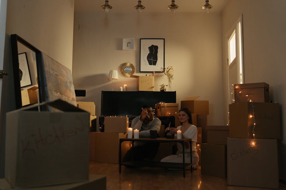 A living room scene during a home relocation process showing two people sitting amidst numerous moving boxes of various sizes, some labeled with handwritten names such as 'clothes' and 'books,' and wrapped with brown packing tape. The boxes are stacked around them, with some placed on the floor and others on furniture. The room features a black flat-screen TV mounted on the wall, a framed minimalist black and white artwork, a small side lamp, and a round decorative mirror with a gold frame. Candle-like lights are arranged on the floor and tables, providing warm ambient lighting. The individuals are engaged with their phones, surrounded by packing materials, and appear to be taking a break during the furniture transport and packing process. The scene reflects careful packing, the importance of organizing goods for a smooth moving day, in alignment with professional removals services by Kentish Town Removals.