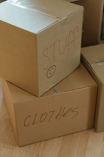 Close-up of two stacked cardboard boxes used for house removal, placed on a wooden floor inside a property. The top box has the word 'STUFF' written in red marker along with a simple smiley face, indicating its contents. The bottom box is labeled 'CLOTHES' in similar handwriting. The boxes are sealed with packing tape and are positioned near other moving boxes, suggesting an ongoing packing process as part of a home relocation. The setting appears well-lit, and the boxes are likely being prepared for transport. This image illustrates typical packing for furniture transport and moving logistics in a house removal, which Kentish Town Removals might facilitate during client home moves.
