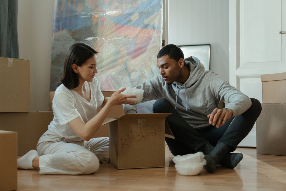 A living room scene during a home relocation process showing two people sitting amidst numerous moving boxes of various sizes, some labeled with handwritten names such as 'clothes' and 'books,' and wrapped with brown packing tape. The boxes are stacked around them, with some placed on the floor and others on furniture. The room features a black flat-screen TV mounted on the wall, a framed minimalist black and white artwork, a small side lamp, and a round decorative mirror with a gold frame. Candle-like lights are arranged on the floor and tables, providing warm ambient lighting. The individuals are engaged with their phones, surrounded by packing materials, and appear to be taking a break during the furniture transport and packing process. The scene reflects careful packing, the importance of organizing goods for a smooth moving day, in alignment with professional removals services by Kentish Town Removals.