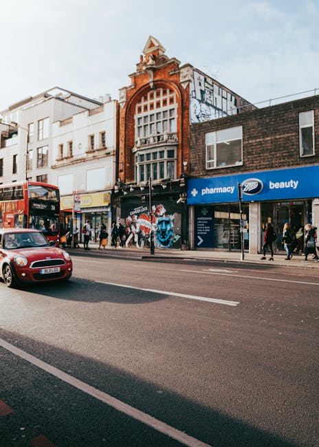 A busy urban street scene in Camden with a row of commercial buildings, including a pharmacy and beauty shop, situated on the ground floor. The buildings feature a mix of architectural styles, with a prominent red brick facade on one structure that has large, arched windows and decorative elements at the roofline. Pedestrians are walking along the pavement in front of the shops, some entering or leaving the stores. A red double-decker bus and a small red car are on the road, which has clearly marked lanes. In the foreground, a loading area or pavement space is visible, where Kentish Town Removals may be arranging furniture, boxes, or packing materials, possibly preparing for a home relocation or furniture transport. The scene is lit with natural daylight, capturing a typical busy day in Camden with activity linked to local moving or delivery services, supporting the context of house removals and logistics.