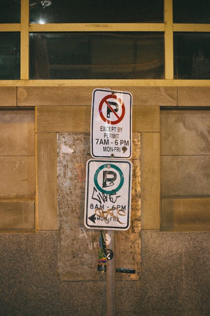 Close-up view of a metal parking sign post attached to a stone wall with two rectangular signs. The top sign indicates no parking except by permit from 7 am to 6 pm, Monday to Friday, with a red circle and slash over a black 'P' symbol. The bottom sign shows a parking icon with a green circle and a 'P' symbol, and specifies parking hours from 8 am to 6 pm, Monday to Friday, with an illustration of hands holding a parking ticket. Above the signs, there is a window with multiple panes reflected in darkness. The signage is part of a vehicle loading and parking regulation area in Camden, relevant to house removals and relocation logistics. The signs are mounted directly to a weathered metal pole, which is positioned in front of a stone facade, indicating restrictions that Kentish Town Removals must observe during home moving activities.