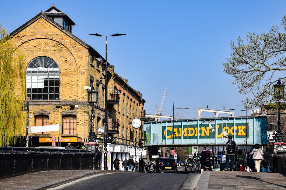 A busy urban street scene in Camden with a row of commercial buildings, including a pharmacy and beauty shop, situated on the ground floor. The buildings feature a mix of architectural styles, with a prominent red brick facade on one structure that has large, arched windows and decorative elements at the roofline. Pedestrians are walking along the pavement in front of the shops, some entering or leaving the stores. A red double-decker bus and a small red car are on the road, which has clearly marked lanes. In the foreground, a loading area or pavement space is visible, where Kentish Town Removals may be arranging furniture, boxes, or packing materials, possibly preparing for a home relocation or furniture transport. The scene is lit with natural daylight, capturing a typical busy day in Camden with activity linked to local moving or delivery services, supporting the context of house removals and logistics.